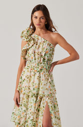 A woman is wearing a sleeveless, one-shoulder dress made from a floral-printed fabric. The dress features a ruffled shoulder detail, an elastic waistband, and a tiered skirt with a high slit.